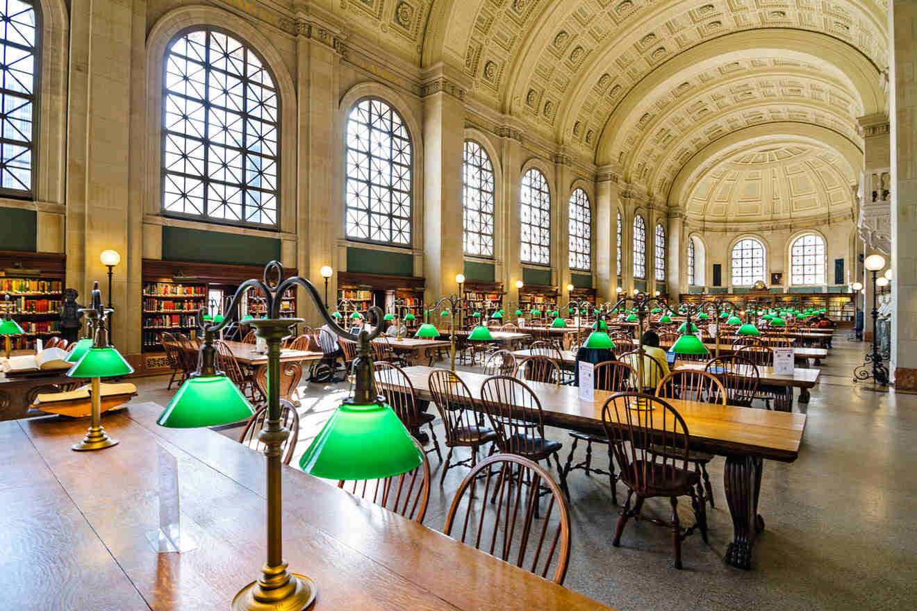 Spacious reading room in the Boston Public Library with arched windows, ornate ceiling, and rows of wooden tables with green-shaded lamps.