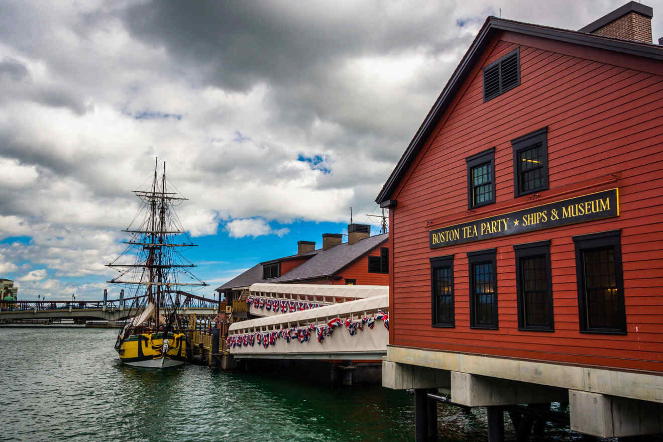 Boston Tea Party Ships & Museum exterior, featuring a historic ship replica docked next to the red-clad museum building with flags under a cloudy sky