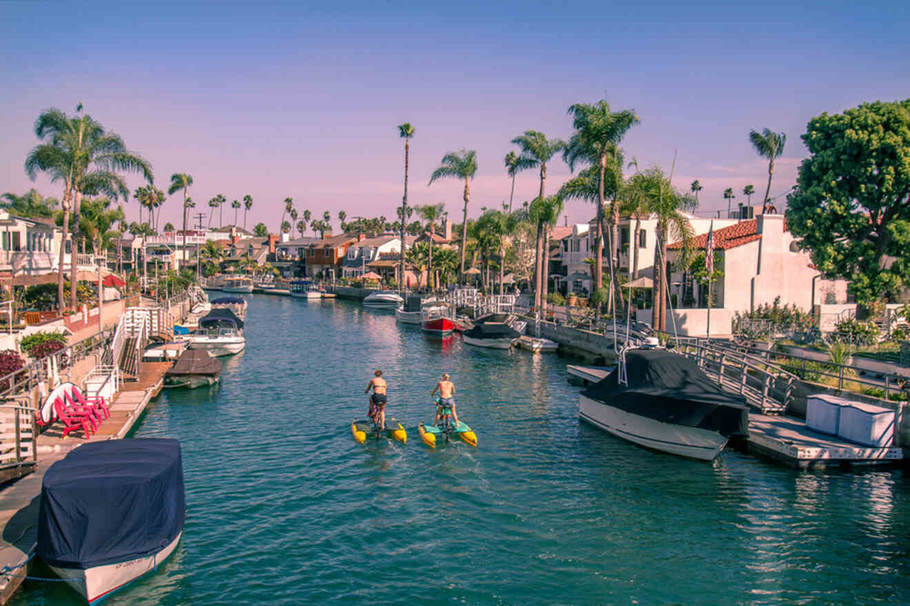 Two people enjoy paddleboarding through the calm waters of a residential canal, with moored boats and palm trees lining the tranquil neighborhood under a clear sky.