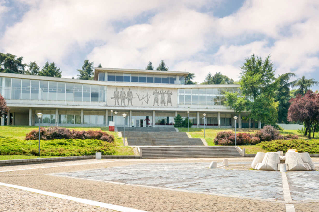 Front view of the Museum of Yugoslavia in Belgrade, a modernist building with a large mosaic depicting historical figures, surrounded by lush greenery