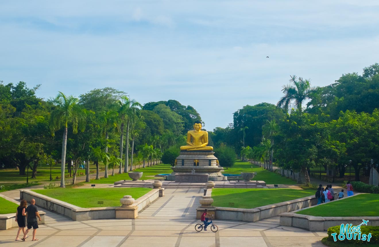 Visitors stroll around a tranquil park with a prominent golden Buddha statue seated atop a pedestal, flanked by lush greenery and palm trees, under a clear blue sky.