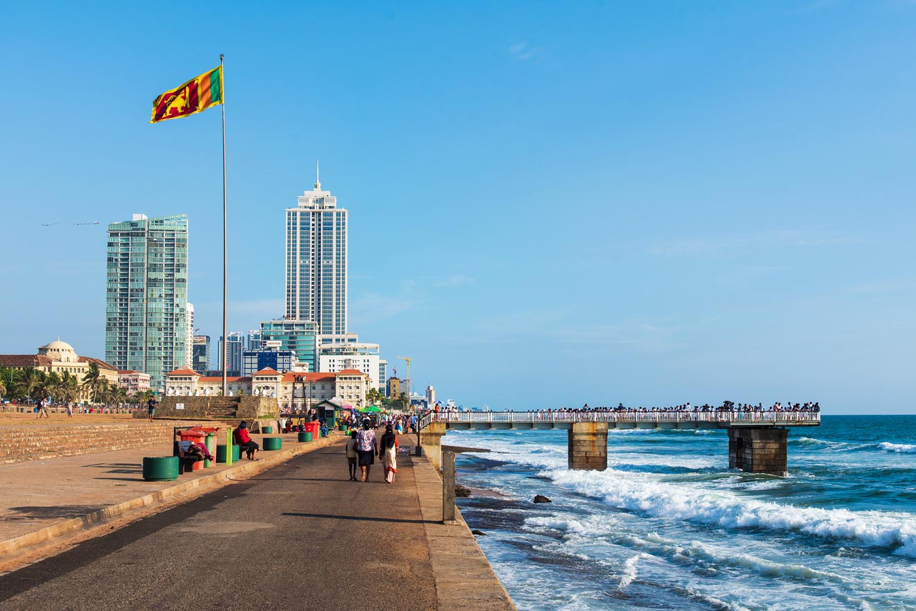 A vibrant scene along a coastal promenade with a large Sri Lankan flag waving, overlooking a bustling pier and modern skyscrapers against a clear blue sky.