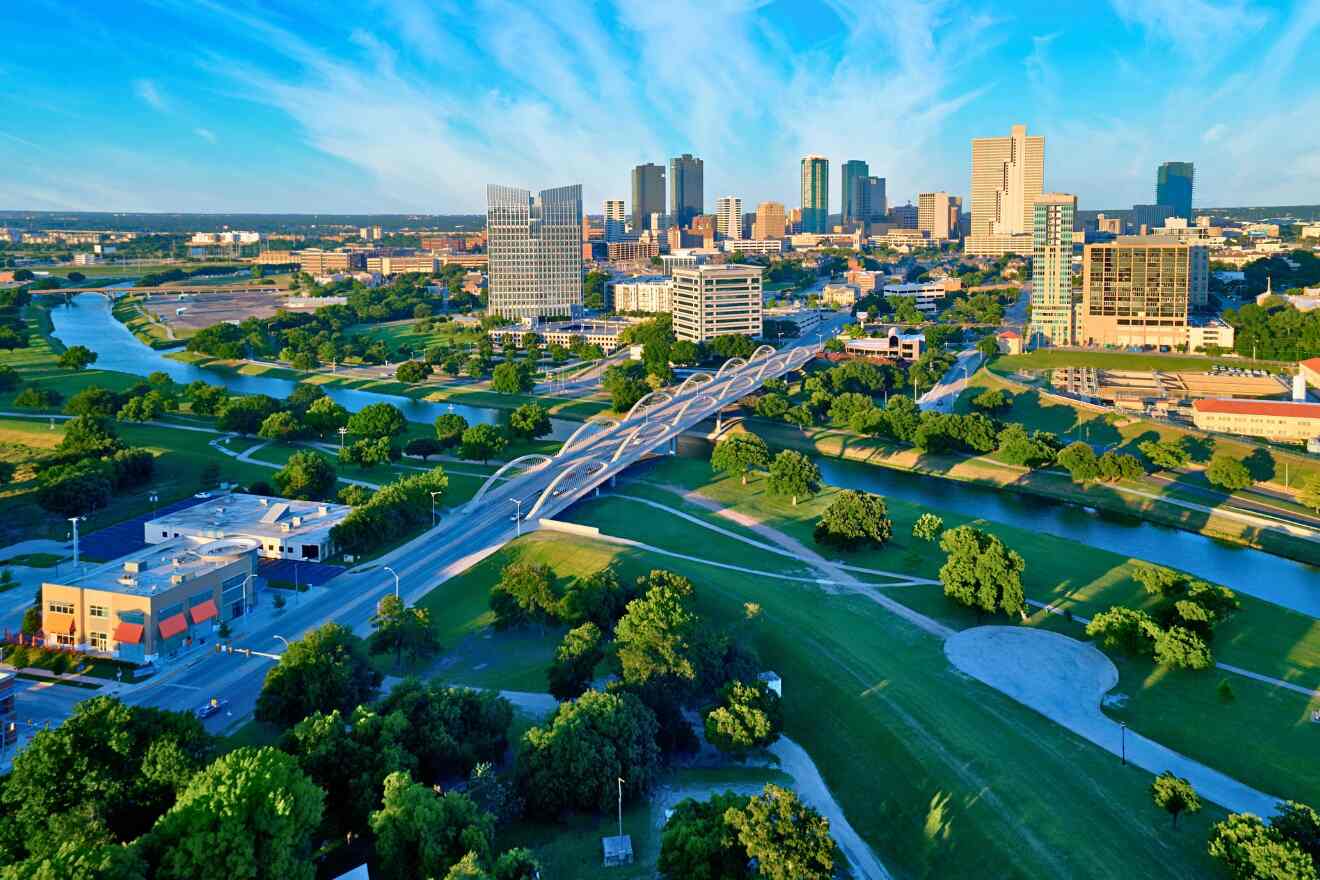 Aerial view of Downtown Forth Worth with a winding river, green spaces, and a modern bridge against a backdrop of high-rise buildings under a clear blue sky.