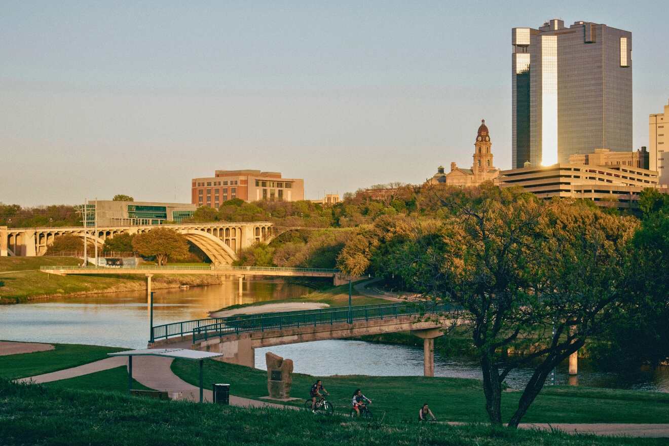 Evening scene of the riverfront in Downtown Forth Worth, showing a bridge with city buildings in the background, and people enjoying the riverside park at sunset.