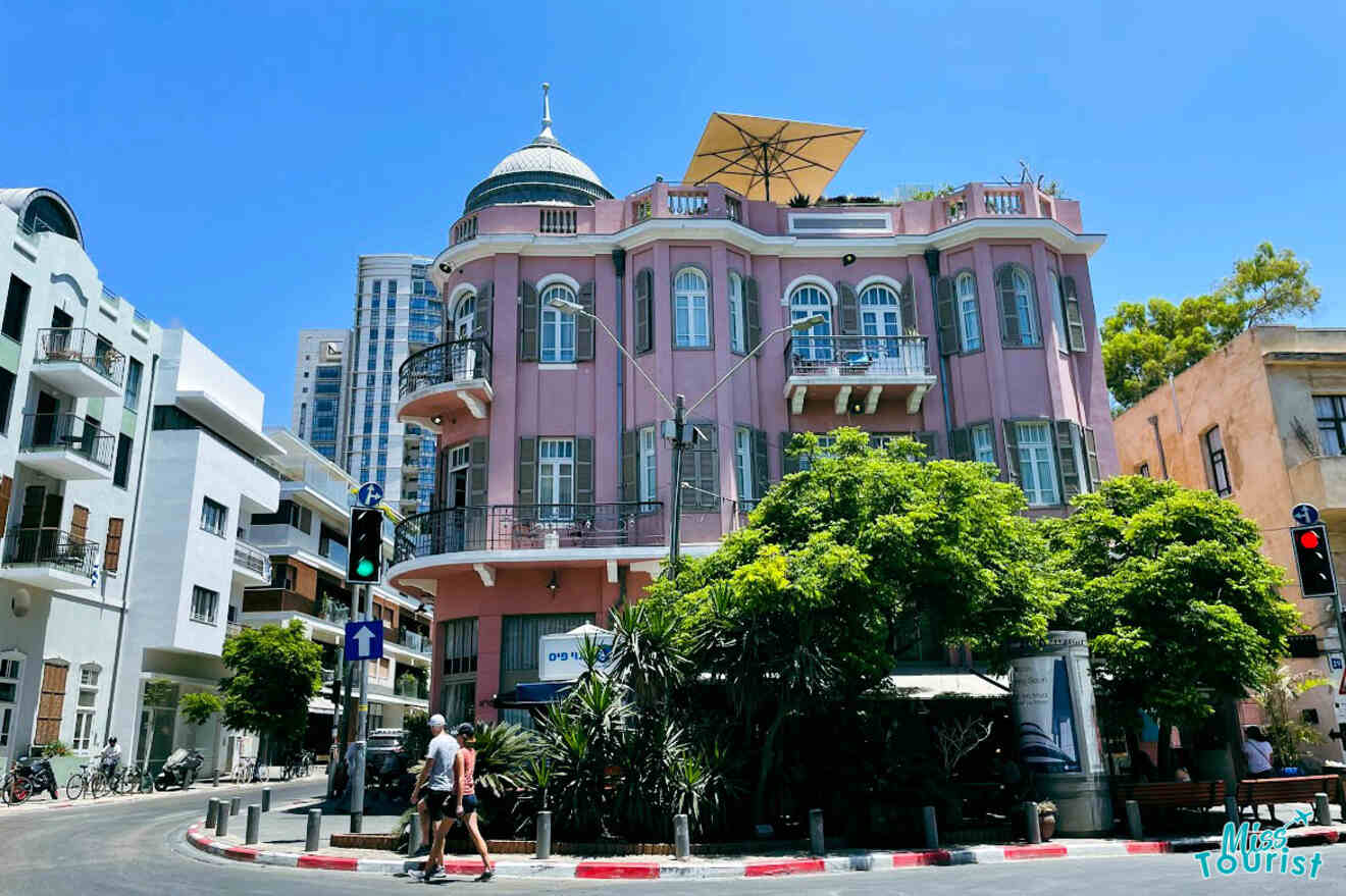 A vibrant corner of Tel Aviv showcasing Bauhaus architecture with a pink building under a clear blue sky, pedestrians crossing the street, and lush greenery surrounding the area