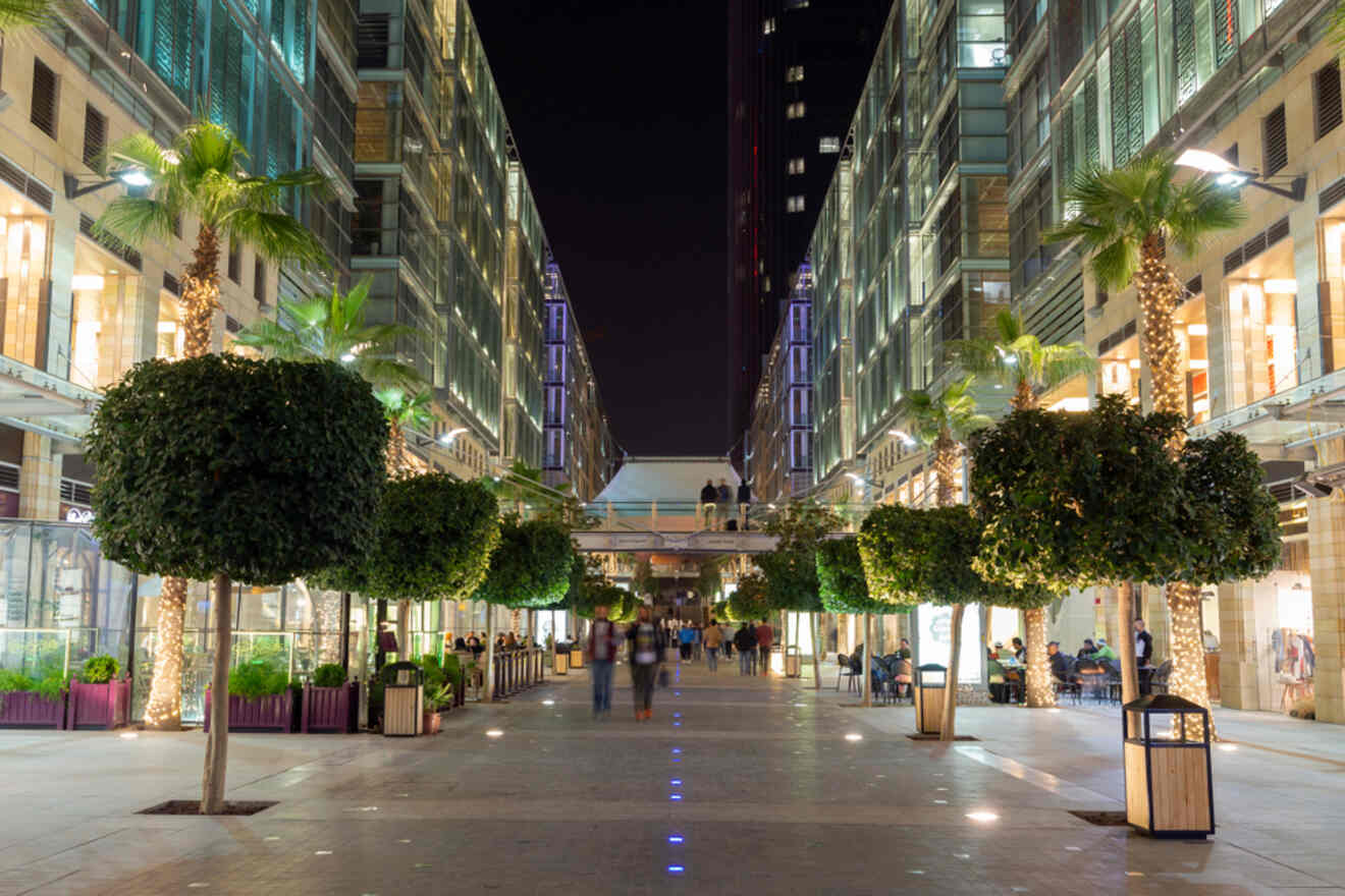 Nighttime scene of Abdali Boulevard in Amman, Jordan, lined with modern buildings, illuminated palm trees, and people strolling along the pedestrian pathway