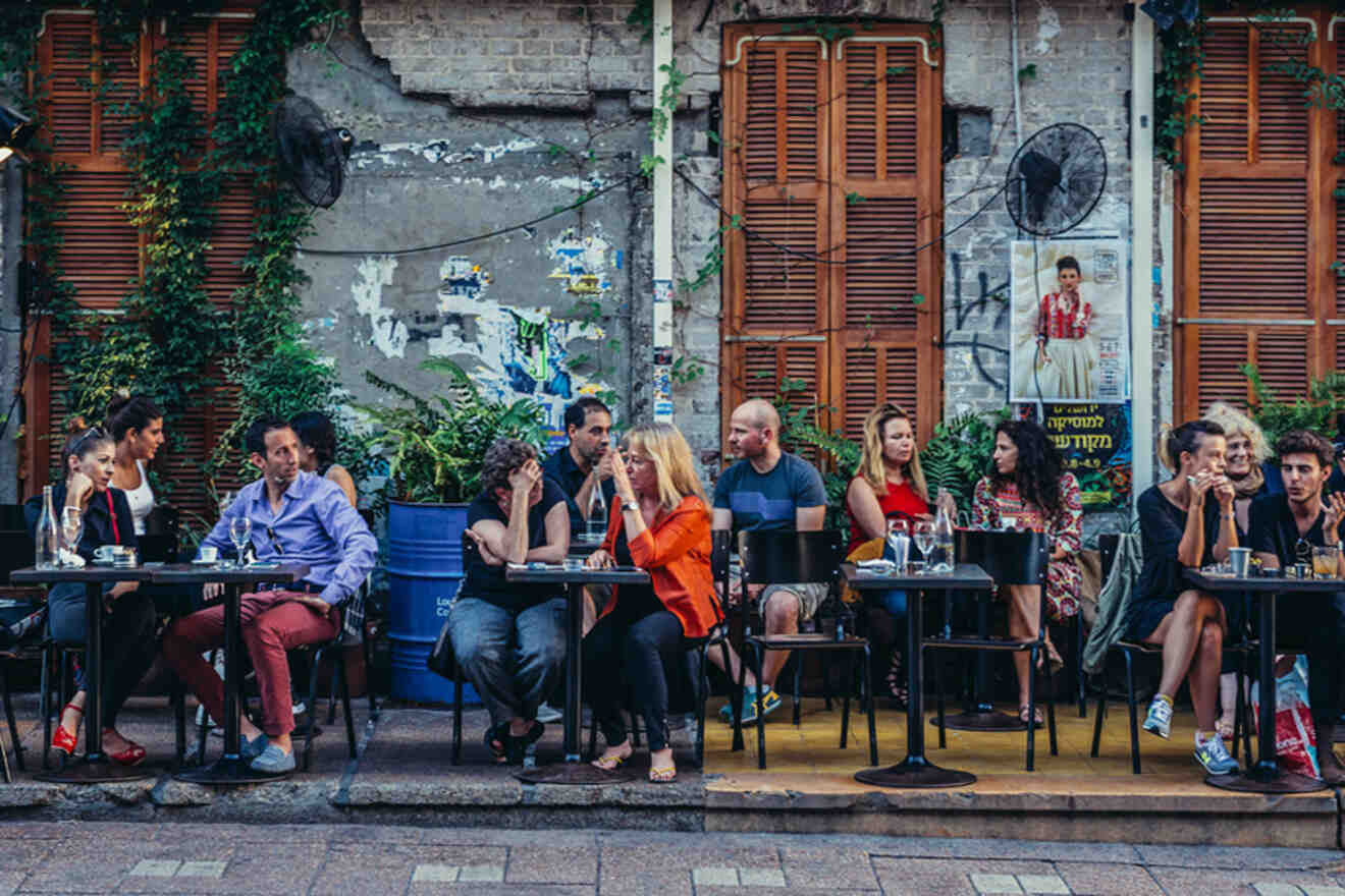 A lively outdoor café scene with patrons enjoying drinks and conversation at tables set along a street with rustic charm and greenery