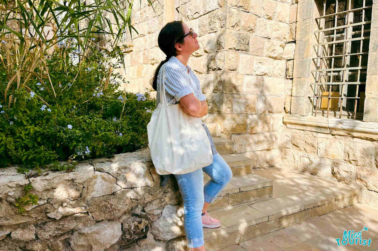 The writer of the post esting against a stone wall in Amman, Jordan, looking contemplatively into the distance, set against a backdrop of lush greenery and traditional architecture