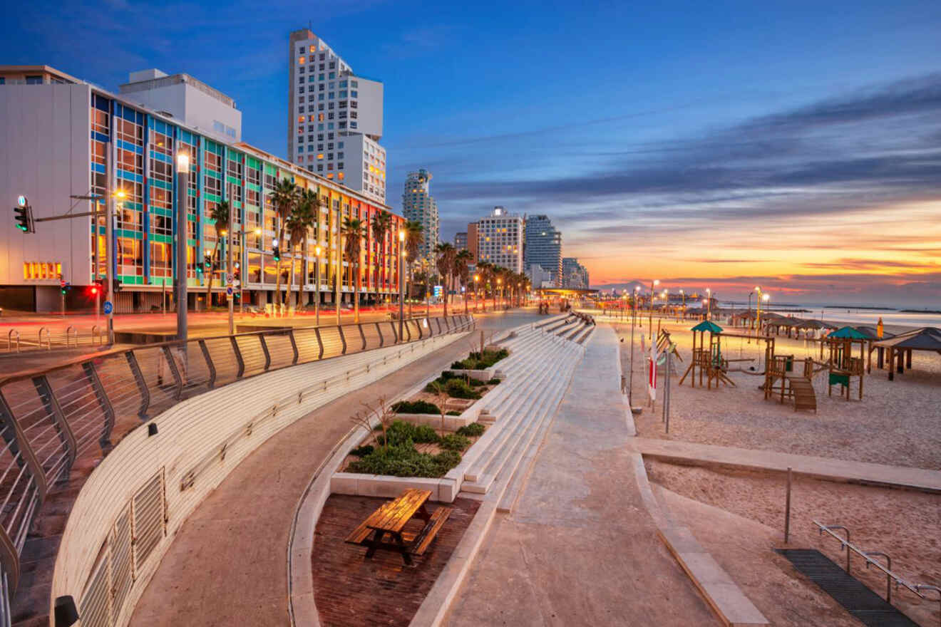 Twilight over the Namal district in Tel Aviv, featuring a modern promenade with palm trees, a sandy beach playground, and a stunning view of the city's coastline and lit buildings