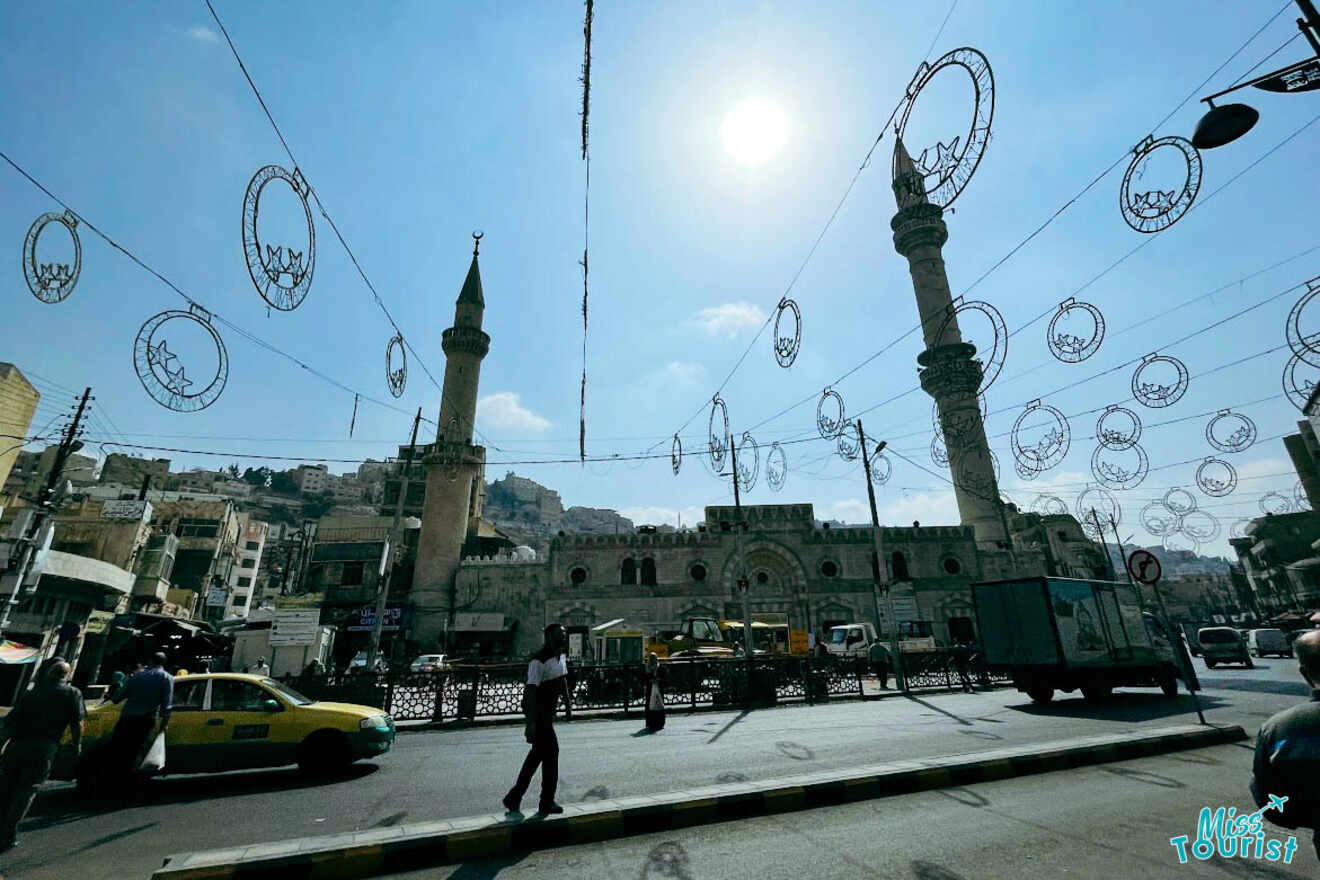 The Grand Husseini Mosque in Amman, Jordan, under a clear sky with decorative street lights and city life bustling around it