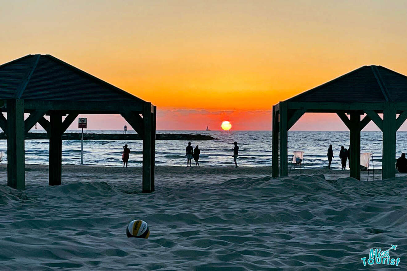 Twilight view of Tel Aviv beach with people gathering under gazebos and a vibrant sunset over the horizon