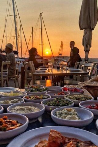 An outdoor dining scene set against a beautiful sunset backdrop, with diners at tables overlooking a harbor with sailboats, and a spread of assorted Mediterranean dishes in the foreground