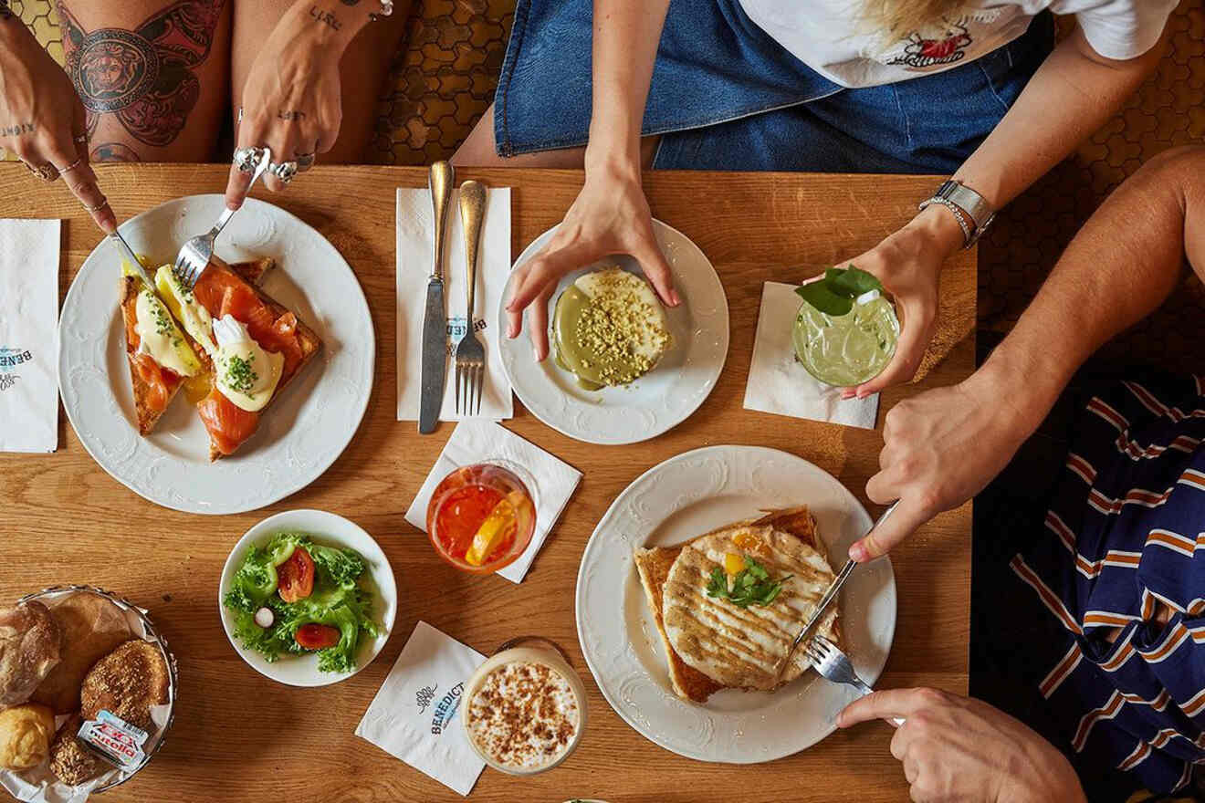 Overhead view of a communal table with hands reaching for assorted breakfast dishes including salmon on toast, grilled sandwiches, and refreshing drinks, depicting a vibrant brunch atmosphere.