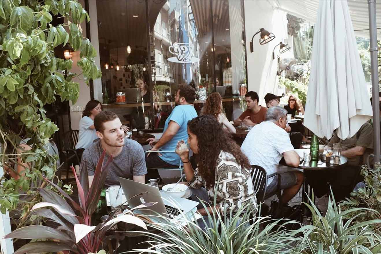 Casual outdoor café setting with patrons eating, working on laptops, and enjoying conversations, surrounded by lush plants and urban architecture reflecting a trendy city vibe