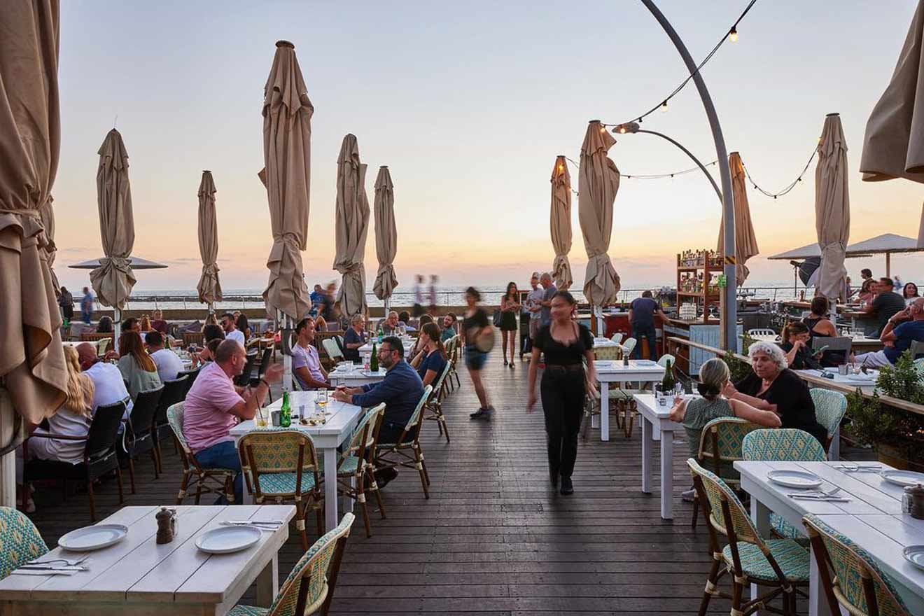 Dusk settling over a sea-view restaurant with diners seated at tables, the ambiance is serene with closed parasols and soft lighting, encapsulating a tranquil dining experience by the waterfront